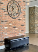 A large industrial-style clock with Roman numerals is mounted on a red brick wall. Below the clock is a black leather bench with a sleek design. The area features an additional wall made of stacked stone tiles, adding texture to the interior.