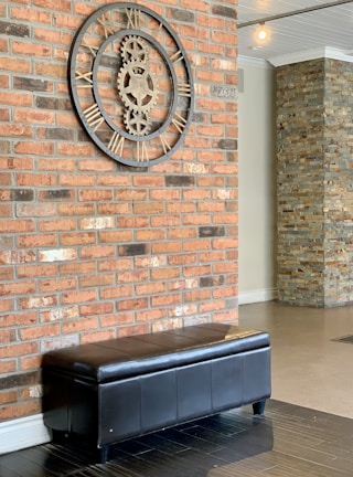 A large industrial-style clock with Roman numerals is mounted on a red brick wall. Below the clock is a black leather bench with a sleek design. The area features an additional wall made of stacked stone tiles, adding texture to the interior.