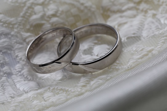 Two silver wedding rings rest on a delicate, embroidered fabric with soft, intricate patterns. The rings have a smooth and polished surface, and their close placement suggests a connection or bond. The background fabric is in shades of white and cream, adding an elegant and sophisticated touch.