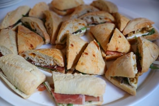 A platter of sandwiches arranged in a circular pattern, each sandwich is cut into triangular pieces. They appear to be filled with a variety of ingredients, including lettuce, cheese, and slices of meat. The bread has a lightly toasted, golden-brown crust.