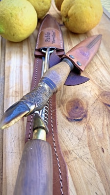 A close-up image of a knife with a textured handle and a metallic blade, resting on a wooden surface. Next to the knife, there are large yellow lemons, and a leather sheath with a name embossed on it.