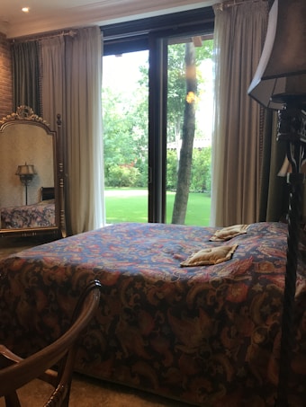 A cozy bedroom with a large bed covered in a richly patterned bedspread, situated in front of a glass door that opens to a lush green garden. A decorative mirror, lamp, and chair are also part of the decor, adding to the vintage feel.