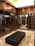 A luxurious walk-in closet featuring dark wooden cabinetry with glass doors, multiple rows of clothes hanging neatly, and a black tufted leather bench in the center. The ceiling is illuminated with recessed lighting, enhancing the elegant and organized appearance of the space. The floor is made of polished stone tiles, adding to the sophisticated ambiance.
