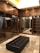 A luxurious walk-in closet featuring dark wooden cabinetry with glass doors, multiple rows of clothes hanging neatly, and a black tufted leather bench in the center. The ceiling is illuminated with recessed lighting, enhancing the elegant and organized appearance of the space. The floor is made of polished stone tiles, adding to the sophisticated ambiance.