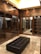 A luxurious walk-in closet featuring dark wooden cabinetry with glass doors, multiple rows of clothes hanging neatly, and a black tufted leather bench in the center. The ceiling is illuminated with recessed lighting, enhancing the elegant and organized appearance of the space. The floor is made of polished stone tiles, adding to the sophisticated ambiance.