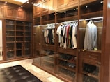 A luxurious walk-in closet with wooden cabinetry and glass doors showcases a neatly organized collection of clothes on hangers. Various shirts and jackets are arranged by color, and shoes are displayed on lower shelves. The warm lighting highlights the richness of the wood.