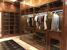 A luxurious walk-in closet with wooden cabinetry and glass doors showcases a neatly organized collection of clothes on hangers. Various shirts and jackets are arranged by color, and shoes are displayed on lower shelves. The warm lighting highlights the richness of the wood.