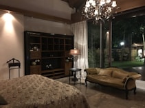 A cozy and elegant room featuring a large window overlooking a garden. The room is decorated with a chandelier, a vintage sofa, and a decorative lamp beside a dark wooden shelf. Natural light from large curtains and soft wall lighting enhances the ambiance.