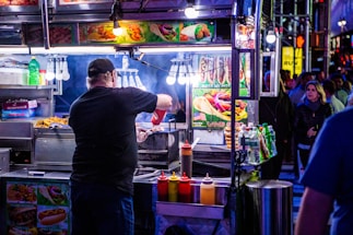 A bustling street food vendor scene at night with a man preparing food at a brightly lit stall. Various condiments are lined up in front of the stall, and colorful pictures of food are displayed above. In the background, people are walking by on the street.