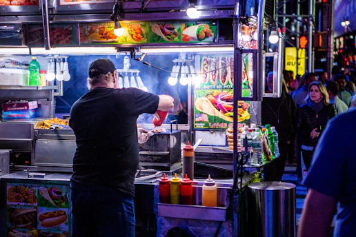 A bustling street food vendor scene at night with a man preparing food at a brightly lit stall. Various condiments are lined up in front of the stall, and colorful pictures of food are displayed above. In the background, people are walking by on the street.