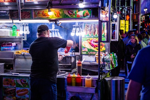 A bustling street food vendor scene at night with a man preparing food at a brightly lit stall. Various condiments are lined up in front of the stall, and colorful pictures of food are displayed above. In the background, people are walking by on the street.