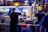 A bustling street food vendor scene at night with a man preparing food at a brightly lit stall. Various condiments are lined up in front of the stall, and colorful pictures of food are displayed above. In the background, people are walking by on the street.
