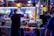A bustling street food vendor scene at night with a man preparing food at a brightly lit stall. Various condiments are lined up in front of the stall, and colorful pictures of food are displayed above. In the background, people are walking by on the street.