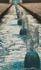 A series of small water fountains are aligned in a straight line, each spouting water upwards. The water appears clear and the spouts are evenly placed along a narrow water feature. The background shows the continuation of this structure with a blurred effect, suggesting depth.