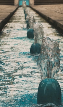 A series of small water fountains are aligned in a straight line, each spouting water upwards. The water appears clear and the spouts are evenly placed along a narrow water feature. The background shows the continuation of this structure with a blurred effect, suggesting depth.