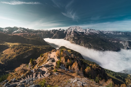 A scenic view of a mountain range with snow-capped peaks under a clear blue sky. A river of clouds flows through the valley, creating a dramatic contrast with the rugged terrain. In the foreground, a trail winds along the ridgeline, surrounded by autumn-colored trees.