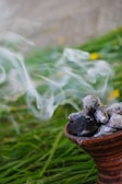 A clay pot containing burning coals with smoke rising from it, set on a bed of green grass with some yellow flowers blurred in the background.