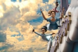 Two animated characters, a young girl and a boy, are flying through a cloudy sky in a whimsical setting. They are dressed in light blue clothes and appear joyful as they soar among fluffy clouds. The sky is vibrant, filled with a mix of bright yellow and soft blue colors, creating a playful and adventurous atmosphere. Large letters can be seen partially in the background, suggesting they are part of a sign or structure.