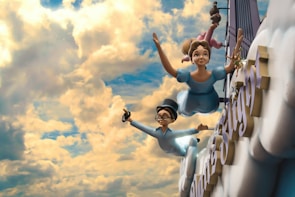 Two animated characters, a young girl and a boy, are flying through a cloudy sky in a whimsical setting. They are dressed in light blue clothes and appear joyful as they soar among fluffy clouds. The sky is vibrant, filled with a mix of bright yellow and soft blue colors, creating a playful and adventurous atmosphere. Large letters can be seen partially in the background, suggesting they are part of a sign or structure.