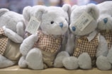 A group of plush teddy bears with blue noses are dressed in checkered brown and cream outfits, sitting closely together on a wooden surface. Each bear has a label attached to its ear, detailing product information.
