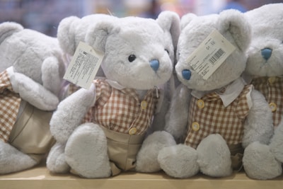 A group of plush teddy bears with blue noses are dressed in checkered brown and cream outfits, sitting closely together on a wooden surface. Each bear has a label attached to its ear, detailing product information.