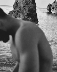 A black and white photograph featuring a rocky shoreline with large formations protruding from calm water. In the foreground, a person is partially visible, emphasizing the contrast with the natural landscape. The ocean stretches out into the horizon.