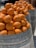 A collection of ripe, bright orange fruits stacked in large, rustic barrels. The sunlight accentuates their vibrant color and smooth texture. The barrels are made of a metal material, and arranged in a line, suggesting abundance and freshness.