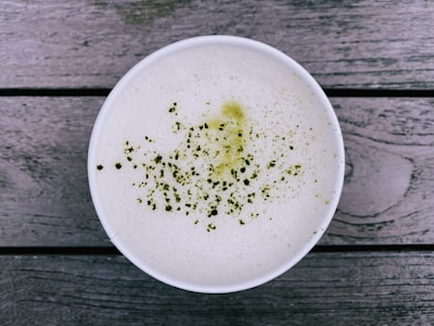 A cup of frothy beverage, likely a matcha latte, is seen from above. The surface is sprinkled with fine green powder, and the cup is placed on a rustic wooden surface with visible texture.