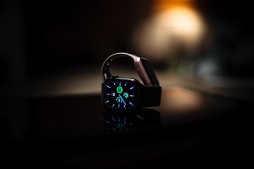 A smartwatch with a lit display showing colorful icons and numbers is placed on a reflective surface. The background is dark with a subtle warm light, possibly from a lamp, creating a contrast with the watch's screen.