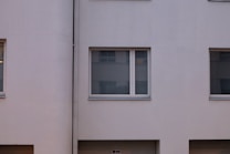 A minimalist facade of a building with smooth, light-colored walls and several large windows that have closed blinds. The design is simple with clean lines and a uniform appearance.