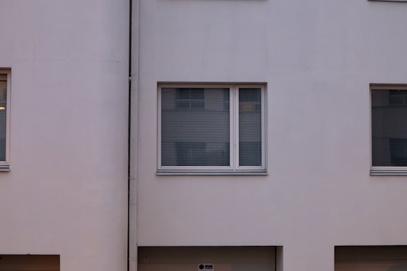 A minimalist facade of a building with smooth, light-colored walls and several large windows that have closed blinds. The design is simple with clean lines and a uniform appearance.