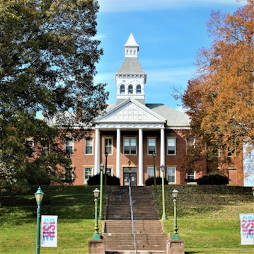 A historic brick building is centered with a grand symmetrical facade. It features white columns supporting a triangular pediment and a distinctive clock tower. An inviting staircase leads up to the entrance, flanked by vintage-style lampposts. Large trees with autumn foliage frame both sides of the scene, enhancing the picturesque setting.