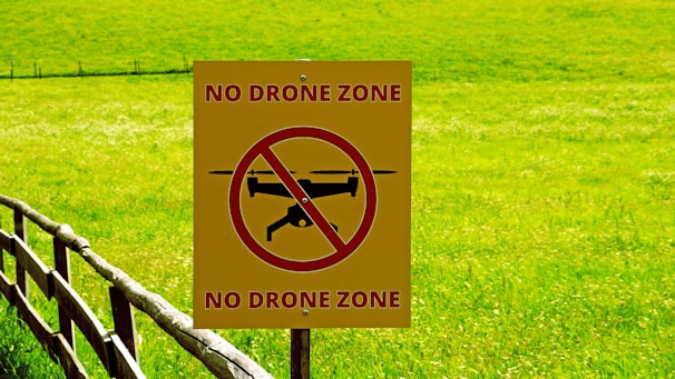 A yellow sign with a red circle and slash over a black drone silhouette stands in front of a lush green field. The words 'NO DRONE ZONE' are written in red at the top and bottom of the sign. A wooden fence runs through the area, adding to the rustic scenery.