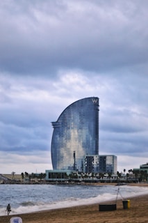 A modern, sail-shaped skyscraper stands beside a beach, reflecting the cloudy sky on its glass facade. The shoreline features gentle waves and a sandy beach, with a person walking along the water's edge. Palm trees line the area near the base of the building, contributing to a coastal setting.