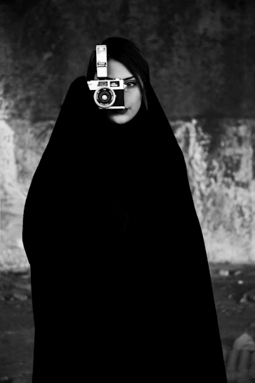 A black and white portrait of a photographer holding a vintage camera against a dark background.