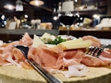 A wooden platter holds slices of cured ham, cheese pieces, and greenery, accompanied by forks. A glass of red wine is in the background, with a blurred, cozy interior setting with warm lighting.