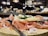 A wooden platter holds slices of cured ham, cheese pieces, and greenery, accompanied by forks. A glass of red wine is in the background, with a blurred, cozy interior setting with warm lighting.