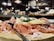 A wooden platter holds slices of cured ham, cheese pieces, and greenery, accompanied by forks. A glass of red wine is in the background, with a blurred, cozy interior setting with warm lighting.