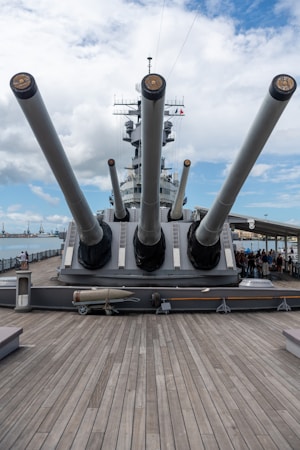 A historical battleship is prominently displayed with large naval gun turrets pointed outward. The wooden deck leads towards the massive guns, and there is a torpedo visible on the deck. In the background, the ship&rsquo;s radar equipment and communication antennas are visible against a partly cloudy sky.