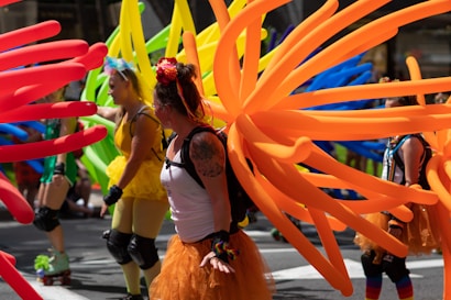 Individuals wearing colorful costumes and elaborate balloon decorations participate in a lively parade. Brightly colored balloons in shades of orange, red, yellow, green, and blue are attached to their outfits, resembling wings or abstract designs. The participants are dressed in festive attire, including tutus and floral headbands, with some on roller skates.