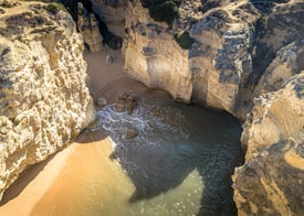 A secluded cove with towering rocky cliffs and a small sandy beach. Waves gently lap against the shore, with sunlight casting dramatic shadows on the sand and water.