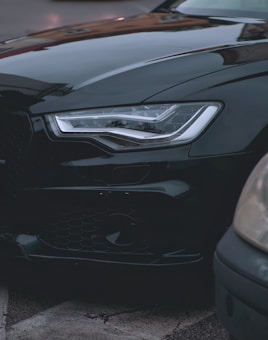 The image displays a close-up view of the front portion of a sleek, black car, focusing on the headlight and part of the bumper. The surroundings suggest an urban environment with the presence of another vehicle partially visible on the right.