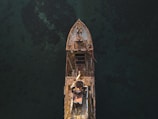 An aerial view of an old, rusted shipwreck partially submerged in dark, clear water. The ship's structure is visibly corroded, with patches of rust and marine growth over its surface. The surrounding water appears calm, and the ship's silhouette is sharply defined against the dark backdrop.