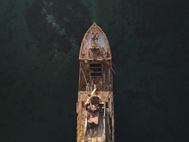 An aerial view of an old, rusted shipwreck partially submerged in dark, clear water. The ship's structure is visibly corroded, with patches of rust and marine growth over its surface. The surrounding water appears calm, and the ship's silhouette is sharply defined against the dark backdrop.