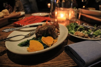 A cozy dining setting features a plate of cooked meat with leafy greens and sliced yellow vegetables on a wooden table. A softly lit candle enhances the warm and inviting atmosphere. A folded napkin sits beside the plate, and other dining items like a partially eaten salad and bread are visible.