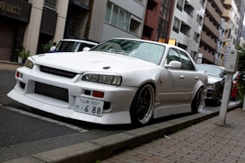 A white sports car is parked on a city street. The car features a streamlined design with large front air intakes and aerodynamic enhancements. It's positioned on a narrow road with a sidewalk paved with rectangular bricks. Surrounding the vehicle are buildings of various heights, featuring a mix of modern and traditional Japanese architecture.