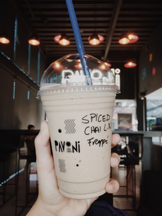 A hand holding a plastic cup with a dome lid and a blue straw. The writing on the cup reads 'SPICED CHAI LATTE Frappe'. The background shows a dimly lit cafe with hanging lights casting a warm glow.