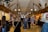 A bustling indoor market hall features numerous stalls lined with various goods, including jewelry, clothing, and souvenirs. The hall has ornately decorated archways and hanging chandeliers, with a high vaulted ceiling. Shoppers, comprising men and women of different ages, are casually browsing and walking through the corridor.