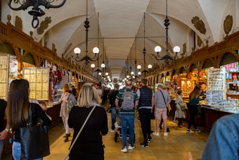 A bustling indoor market hall features numerous stalls lined with various goods, including jewelry, clothing, and souvenirs. The hall has ornately decorated archways and hanging chandeliers, with a high vaulted ceiling. Shoppers, comprising men and women of different ages, are casually browsing and walking through the corridor.