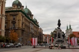 An urban scene featuring a prominent historical monument in a square, surrounded by architectural buildings with detailed facades and domed roofs. Several flags in red and white are displayed around the monument, hinting at a national significance. People are walking and cars are parked along the road, suggesting an active city environment.